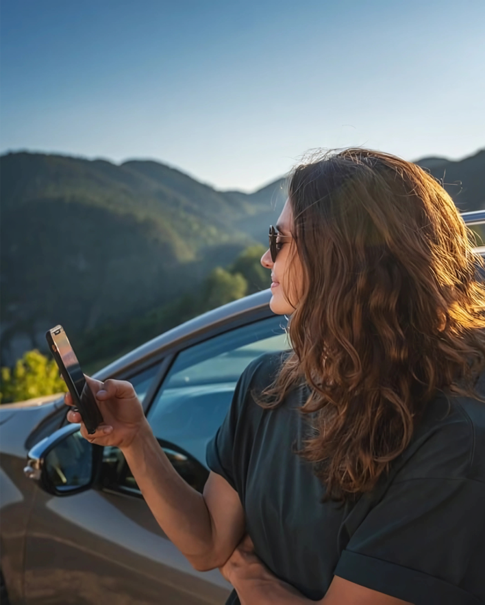imagen de una chica viendo el movil y que tiene como fondo unas montañas y un coche que ha alquilado a través del servicio de alquiler de coche en asturias alvemaco rent